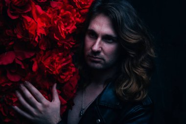 acımasız adam uzun saçlı çıplak bir vücut üzerinde deri ceketli kırmızı çiçek çiçekli dekor yakın duruyor. üzgün ve umutsuz bir yalnız adam