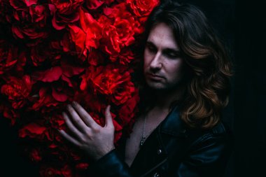 acımasız adam uzun saçlı çıplak bir vücut üzerinde deri ceketli kırmızı çiçek çiçekli dekor yakın duruyor. üzgün ve umutsuz bir yalnız adam