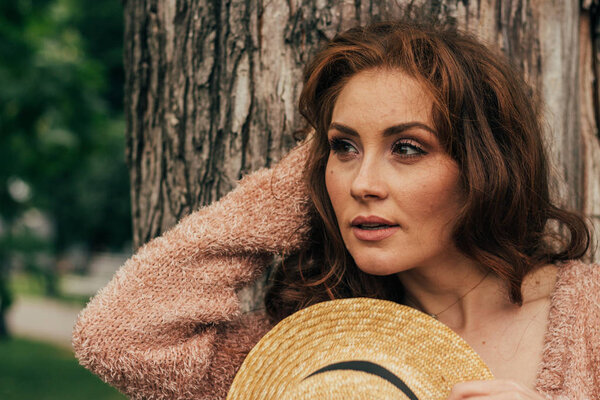portrait of a beautiful girl who stands near a tree. summer, walk in nature, red-haired girl in a vintage dress and a beige sweater. holds a hat, flirts,modesty romance and flirt