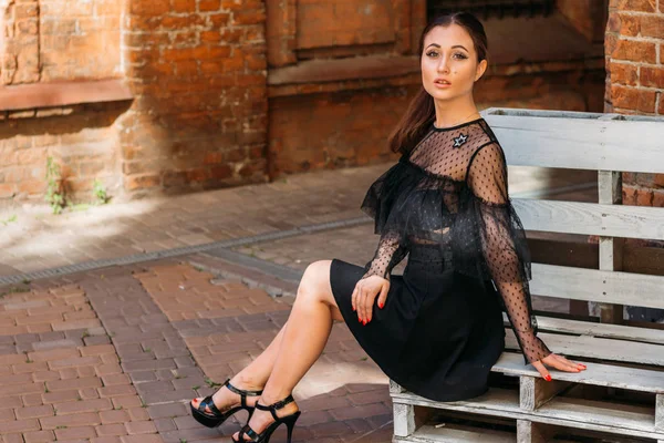 güzel esmer beyaz bir bankta oturan, uzun bacaklarını koyarak, poz, flört, dinleniyor. bir tuğla bina arka plan üzerinde