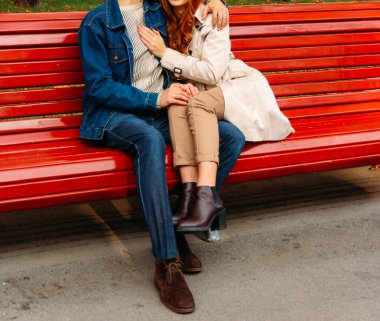 Çift bir bankta oturan bir adam, bir adam bir kadına sarılma bir kız bacaklar. bir adam kot pantolon ve bir kot ceket, bir kız bej bir palto ve pantolon, kahverengi Ayakkabı