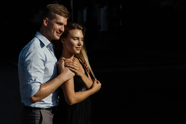 man hugging girl on the street, hugging her, attracted to each other. girl in a black dress. man hugs from behind the woman and kisses on the forehead.the couple is standing against a dark background