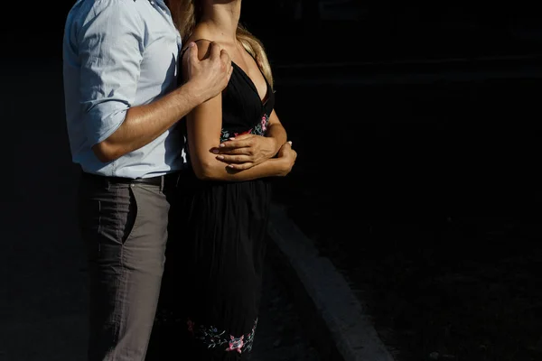 Uomo abbraccia ragazza da dietro, per strada, abbracciandola, attratto l'uno dall'altro. ragazza con un vestito nero. uomo abbraccia da dietro la donna e si bacia sulla fronte. La coppia è in piedi contro un buio — Foto Stock