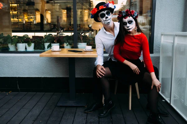 A couple, wearing skull make-up for. All souls day. Boy and girl sugar skull makeup.painted for halloween sit in a cafe. dead in the city. zombie walk.day of the dead holiday in mexico