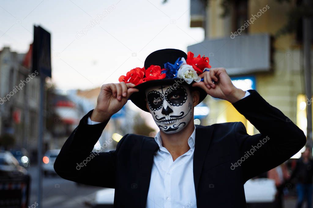 a man with a painted face of a skeleton, a dead zombie, in the city during the day. day of all souls, day of the dead, halloween, ghost walk in a hat