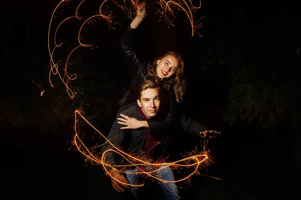 night, a girl in a black jacket jumped on his back to the guy and the couple waving lights on a black background, draws light. holiday, sparklers