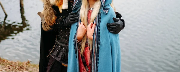 A girl in a blue cloak and a man in ancient clothes, uniforms, protection. stand near the river, hugs, couple, attitude. cosplay of thrones. wolfskin cape. outfit — Stock Photo, Image