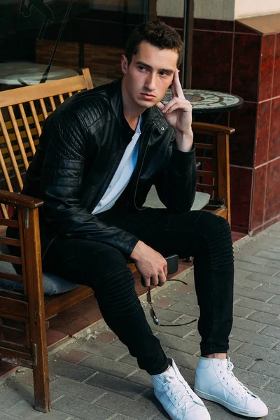 attractive guy sitting on a bench in a cafe, holds sunglasses in his hand, on the street, white sneakers, white T-shirt, black leather jacket and black jeans, pants. model. waiting for a meeting.