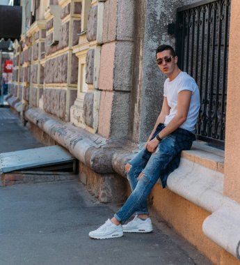 Çekici, seksi bir adam üzerinde bir çıkıntı güneş gözlüğü sokakta duvarındaki bir kaldırıma yaslanmış oturuyor. kendine güven, şişkinlik ve arrogance.model, poz, Genç bir adam portresi