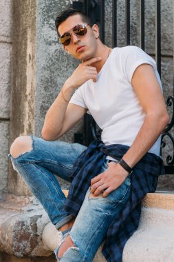 Çekici, seksi bir adam üzerinde bir çıkıntı güneş gözlüğü sokakta duvarındaki bir kaldırıma yaslanmış oturuyor. kendine güven, şişkinlik ve arrogance.model, poz, Genç bir adam portresi