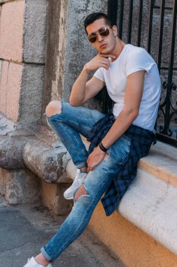 Çekici, seksi bir adam üzerinde bir çıkıntı güneş gözlüğü sokakta duvarındaki bir kaldırıma yaslanmış oturuyor. kendine güven, şişkinlik ve arrogance.model, poz, Genç bir adam portresi