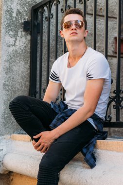 Çekici, seksi bir adam üzerinde bir çıkıntı güneş gözlüğü sokakta duvarındaki bir kaldırıma yaslanmış oturuyor. kendine güven, şişkinlik ve arrogance.model, poz, beyaz spor ayakkabı içinde genç bir adam portresi