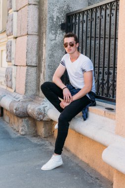 Çekici, seksi bir adam üzerinde bir çıkıntı güneş gözlüğü sokakta duvarındaki bir kaldırıma yaslanmış oturuyor. kendine güven, şişkinlik ve arrogance.model, poz, beyaz spor ayakkabı içinde genç bir adam portresi