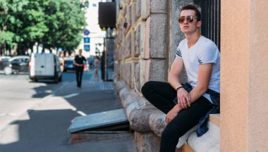 Çekici, seksi bir adam üzerinde bir çıkıntı güneş gözlüğü sokakta duvarındaki bir kaldırıma yaslanmış oturuyor. kendine güven, şişkinlik ve arrogance.model, poz, beyaz spor ayakkabı içinde genç bir adam portresi