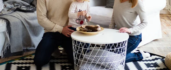 the family sits at the table and makes crafts, is going to decorate the house, on the table are wooden bars, a wooden cut, a log house, a Christmas tree toy,cones.