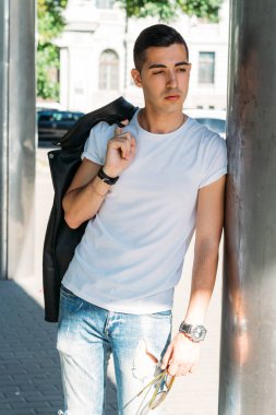 Genç, yakışıklı bir beyaz gömlek, siyah, deri ceket ve mavi, yırtık kot biri sokak, modeli, şehirde yürümek, canlı Style.in'ın beyaz spor ayakkabı. bir saat el, parlak güneş parlar, yaz