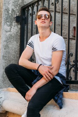 Çekici, seksi bir adam üzerinde bir çıkıntı güneş gözlüğü sokakta duvarındaki bir kaldırıma yaslanmış oturuyor. kendine güven, şişkinlik ve arrogance.model, poz, beyaz spor ayakkabı içinde genç bir adam portresi