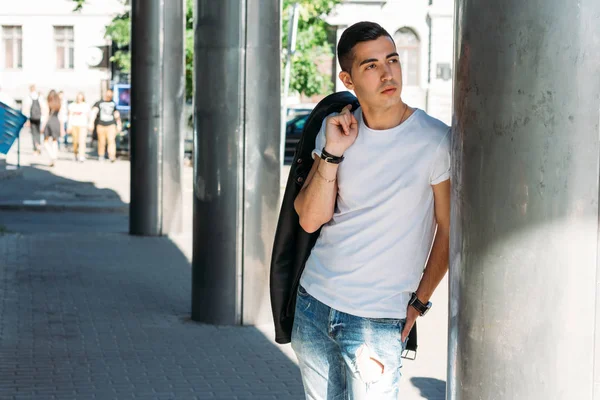 young, handsome guy in a white T-shirt, black, leather jacket and blue, torn jeans is on the street, model, city walk, live style.in white sneakers. with a clock on hand, bright sun shines, summer
