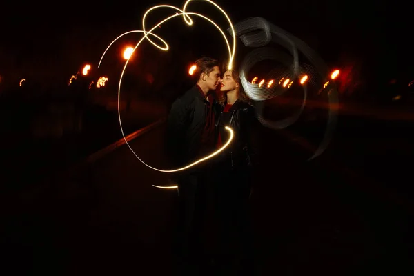 night, a girl in a black jacket jumped on his back to the guy and the couple waving lights on a black background, draws light. holiday, sparklers