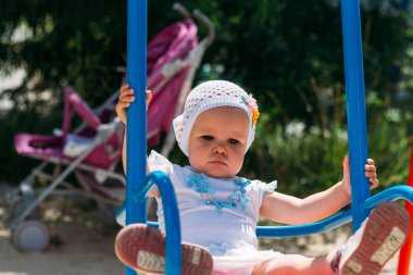 küçük bir kız bir kızın kollarında oturur, o onun arkasından sarıldı ve hands.little kız beyaz elbise ve şapka salıncakta sürme holding, yaz güneş ve ısı. Bahçesi. çocukluk serenity