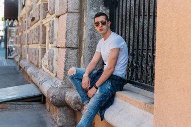 Çekici, seksi bir adam üzerinde bir çıkıntı güneş gözlüğü sokakta duvarındaki bir kaldırıma yaslanmış oturuyor. kendine güven, şişkinlik ve arrogance.model, poz, Genç bir adam portresi