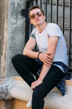 Çekici, seksi bir adam üzerinde bir çıkıntı güneş gözlüğü sokakta duvarındaki bir kaldırıma yaslanmış oturuyor. kendine güven, şişkinlik ve arrogance.model, poz, beyaz spor ayakkabı içinde genç bir adam portresi
