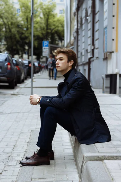 portrait of a man, on the street, in a suit and in a coat. sits on the sidewalk. waiting on the hand wearing a watch.