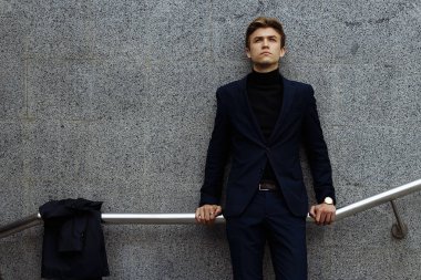 sokakta bir kat asılı, hakkında bir korkuluk, koyu mavi bir takım elbise, yanında, bir korkuluk üzerinde gri bir duvara yaslanmış bir adam duruyor. üzüntü ve dalgınlık. üzgün adam