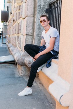 Çekici, seksi bir adam üzerinde bir çıkıntı güneş gözlüğü sokakta duvarındaki bir kaldırıma yaslanmış oturuyor. kendine güven, şişkinlik ve arrogance.model, poz, beyaz spor ayakkabı içinde genç bir adam portresi