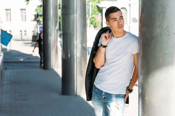 young, handsome guy in a white T-shirt, black, leather jacket and blue, torn jeans is on the street, model, city walk, live style.in white sneakers. with a clock on hand, bright sun shines, summer
