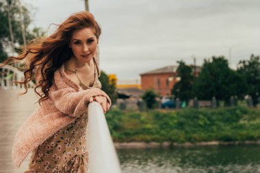 güzel kız köprüde duruyor, korkuluk için tutar ve kameraya doğru görünüyor. rüzgar saçlarını geliştirir. nehir akar aşağıda. kız flört ediyor. aramalar ile