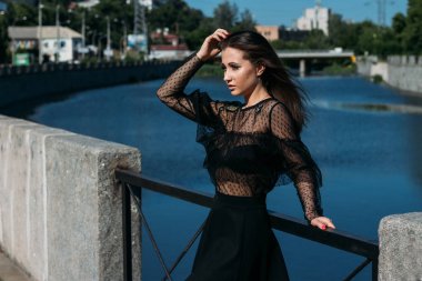 güzel esmer şehrin Nehri yakınında köprünün üzerinde duruyor. siyah elbiseli bir kız bir yüz güneş ve ona esen rüzgar koyar