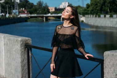 güzel esmer şehrin Nehri yakınında köprünün üzerinde duruyor. siyah elbiseli bir kız bir yüz güneş ve ona esen rüzgar koyar