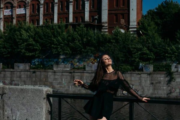 güzel esmer şehrin Nehri yakınında köprünün üzerinde duruyor. siyah elbiseli bir kız bir yüz güneş ve ona esen rüzgar koyar