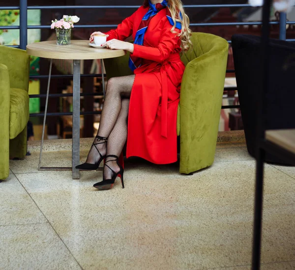 Дівчина в червоному плащі в ажурних колготках, сидить в кафе над чашкою кави, поруч з нею червона парасолька чекає на зустріч — стокове фото
