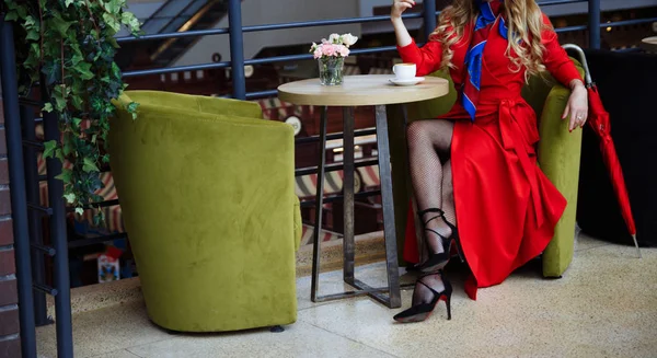 Girl in a red raincoat in fishnet tights, sits in a cafe over a cup of coffee, next to it there is a red umbrella waiting for a meeting — Stock Photo, Image