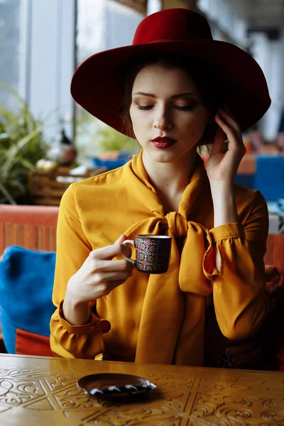 Портрет дівчини в кафе з чашкою кави та капелюхом. портрет чуттєвої молодої дівчини в капелюсі та блузці з бантом. Красива брюнетка в кафе тримає чашку кави — стокове фото