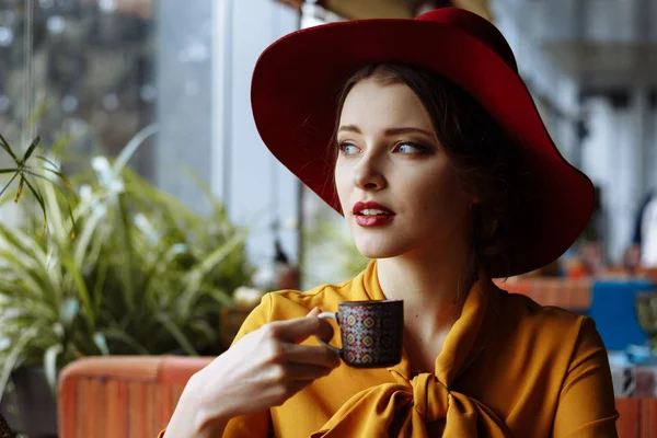 portrait of a girl in a cafe with a cup of coffee and a hat.portrait of sensual young girl wearing floppy hat and blouse with bow. Beautiful brunette woman in cafe holding cup of coffee
