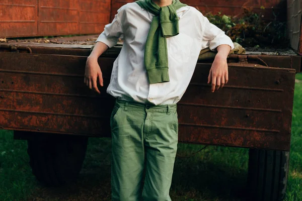 Un ragazzo con una camicia bianca e pantaloni verdi, un maglione verde intorno al collo, appoggiato i gomiti sul vecchio cugino camion, scalzo sull'erba in estate. villaggio, contadino, in fattoria. mani in tasca — Foto Stock
