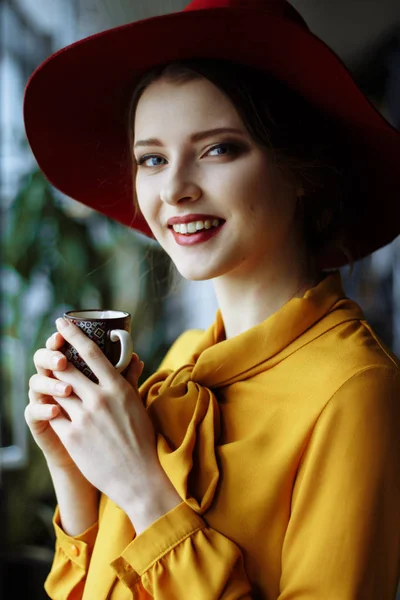 portrait of a girl in a cafe with a cup of coffee and a hat.portrait of sensual young girl wearing hat and blouse with bow. Beautiful brunette woman in cafe holding cup of coffee
