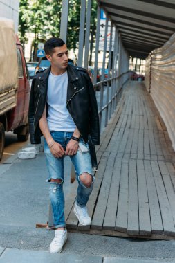 yakışıklı, çekici bir adam, ayakta beyaz bir T-shirt, Korkuluk üzerine eğilerek siyah, deri ceket, siyah pantolon ve beyaz spor ayakkabı, mokasen, sketchboards,