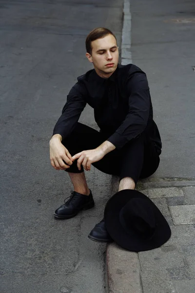 Un uomo in nero con un cappello nero è seduto sull'asfalto sulla strada, sulla strada . — Foto Stock