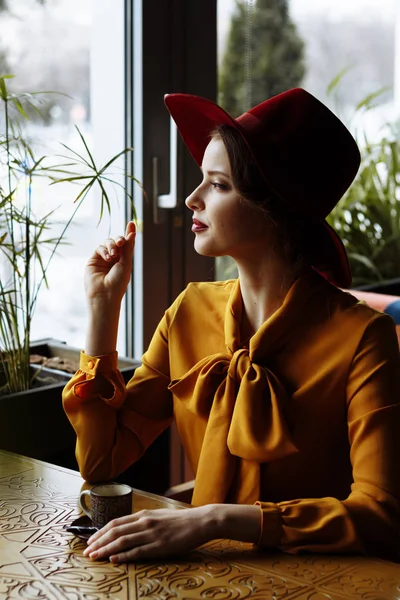 girl in a cafe with a cup of coffee and a hat.portrait of sensual young girl wearing floppy hat and blouse with bow. Beautiful brunette woman in cafe holding cup of coffee