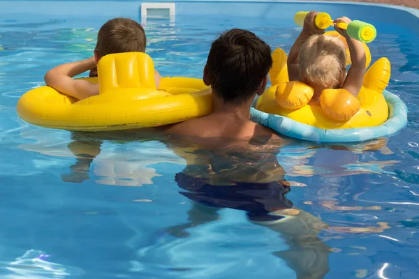 Δύο παιδιά με έναν προπονητή, με τον μπαμπά μαθαίνουν να κολυμπούν στην πισίνα, πίσω όψη. νοιάζεται, διακοπές. φουσκωτά κύκλους. Σωσίβιο — Φωτογραφία Αρχείου