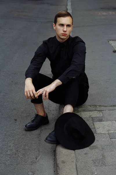 Un uomo in nero con un cappello nero è seduto sull'asfalto sulla strada, sulla strada . — Foto Stock