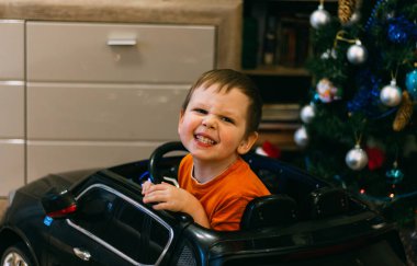 A child is sitting in a toy car, near the Christmas tree. A gift for the new year. grimace rejoices