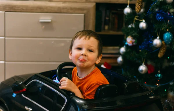 Uma criança está sentada em um carro de brinquedo, perto da árvore de Natal. Um presente para o ano novo. grimace alegra-se — Fotografia de Stock