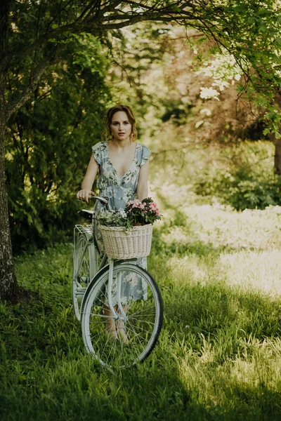 Portret pięknej dziewczyny w lesie, trzymając rower z koszem kwiatów, za promieniami słońca, niebieską kwiecistką sukienkę, letni spacer — Zdjęcie stockowe