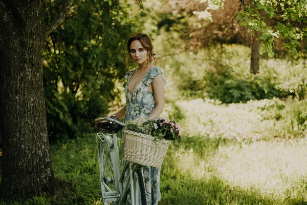 Porträtt av en vacker flicka i skogen, som håller en cykel med en korg med blommor, bakom solens strålar, en Blå blommig klänning, sommar promenad — Stockfoto