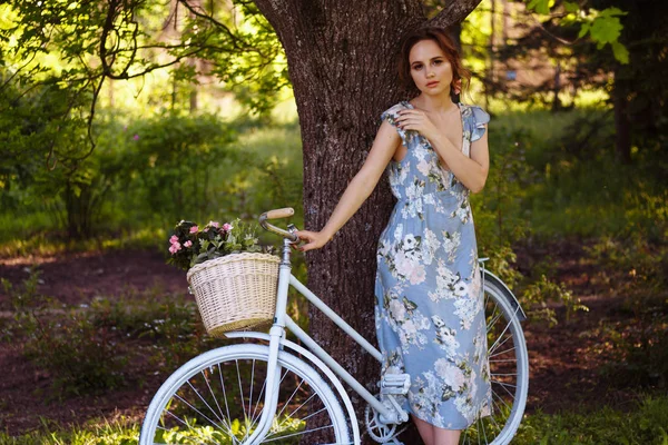 Porträtt av en vacker flicka i skogen, som håller en cykel med en korg med blommor, bakom solens strålar, en Blå blommig klänning, sommar promenad — Stockfoto